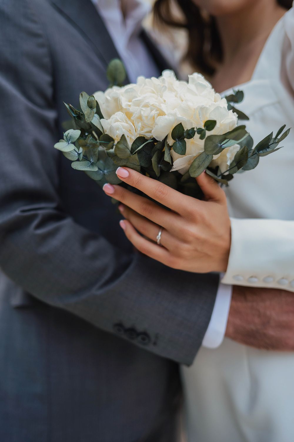 A wedding bouquet of white peonies and eucalyptus leaves held by hands wearing formal attire and a wedding ring.