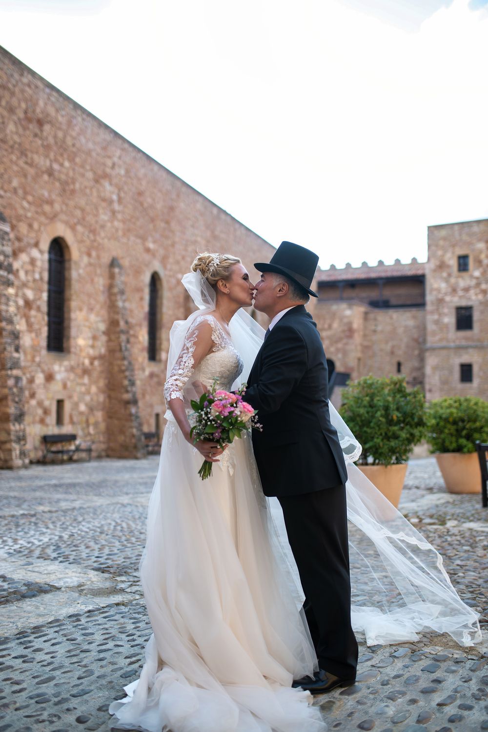 Romantic wedding couple sharing a kiss at historic stone castle courtyard with cobblestone ground and flowing veil.