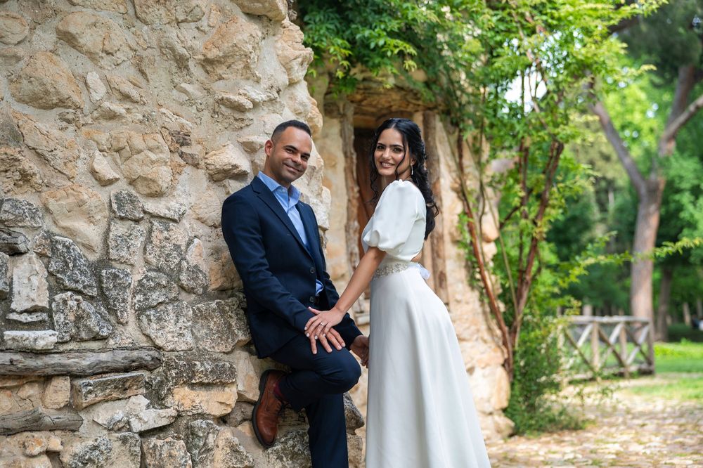 A couple holding hands and posing against an old stone wall in a romantic garden setting.