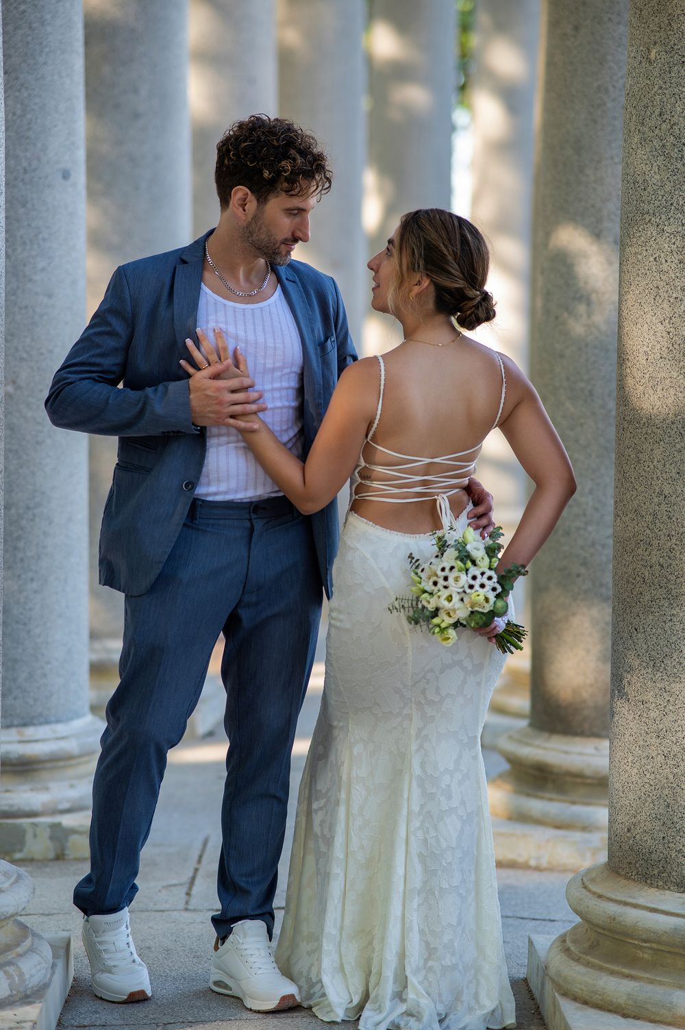 Bride in backless lace gown holds bouquet while sharing intimate moment with groom in navy suit near stone columns.
