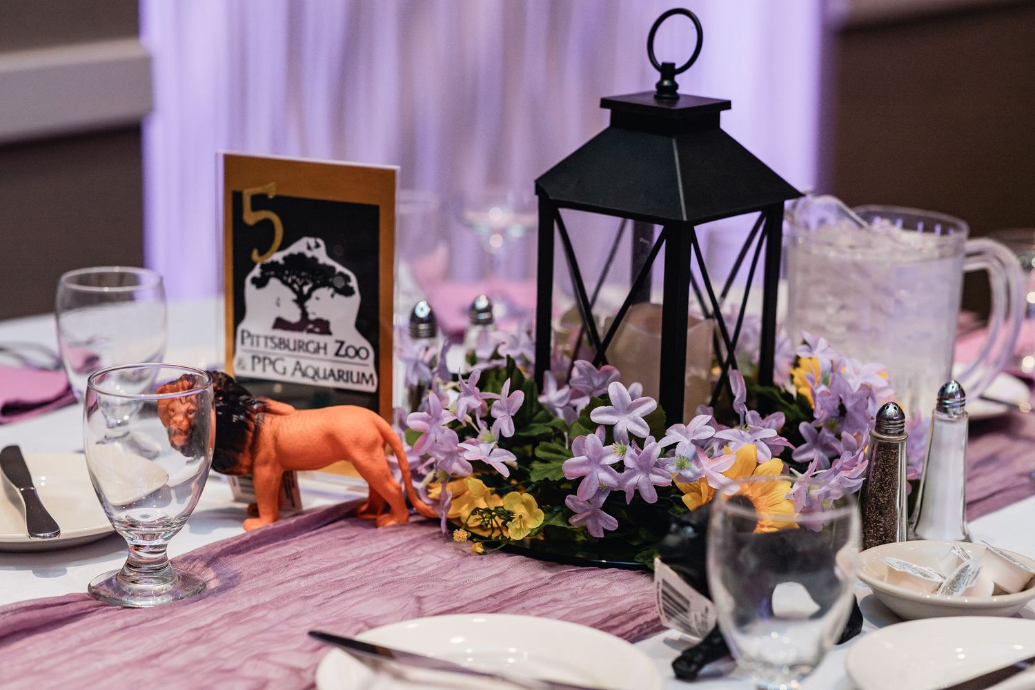 A table setting with a black lantern centerpiece, purple flowers, and framed photo amid rustic wedding decor details.