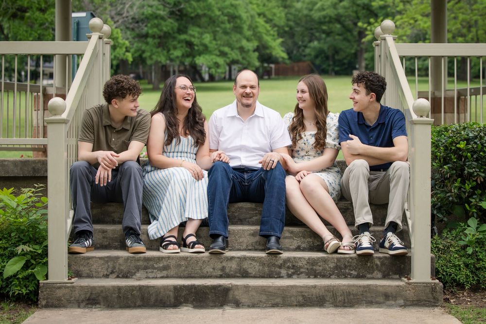A group relaxes together on stone steps surrounded by greenery and a wooden railing on a sunny day.