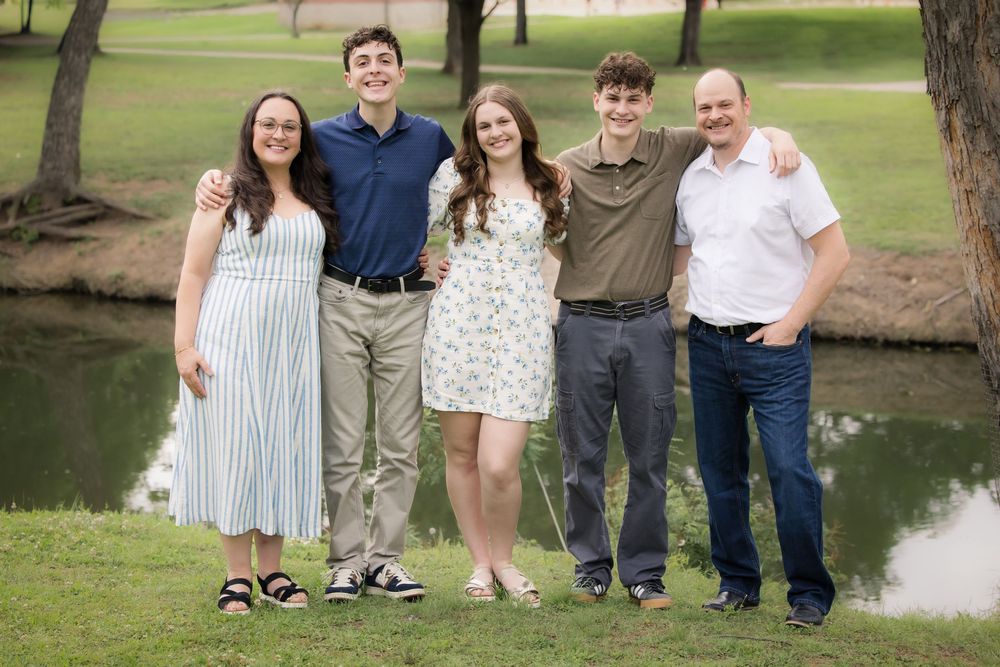 A group of five people stands together outdoors near a pond in a park setting on a sunny day.