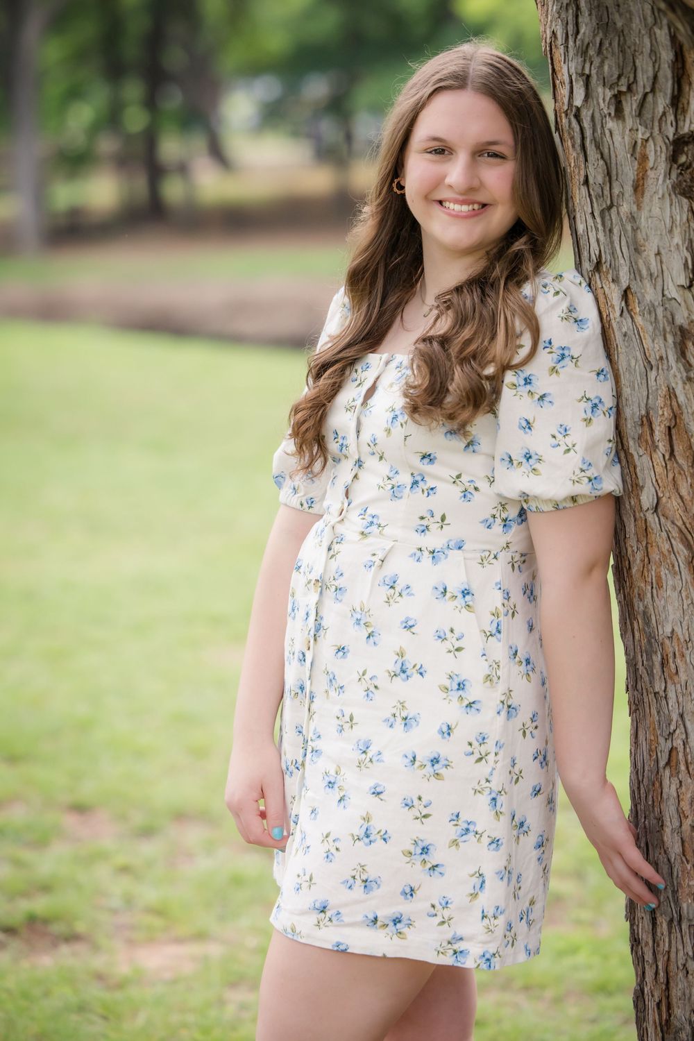 A person in a white floral dress stands next to a tree trunk in a park setting with green grass in the background.