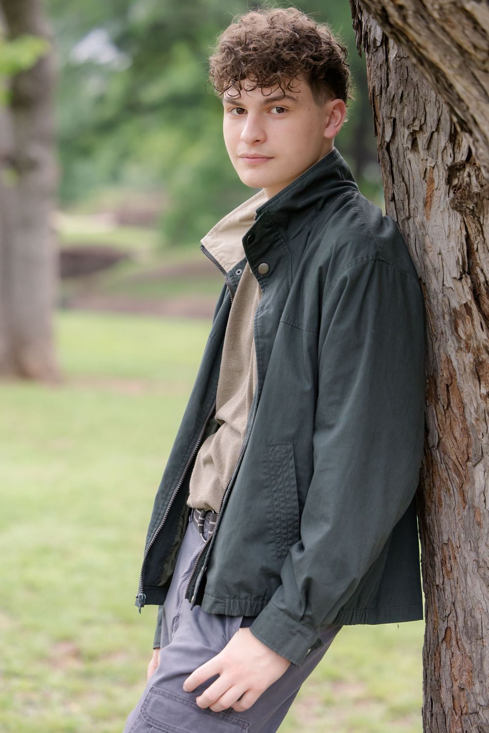 Person wearing a black jacket and gray pants leans against a tree trunk in an outdoor park setting with blurred greenery.
