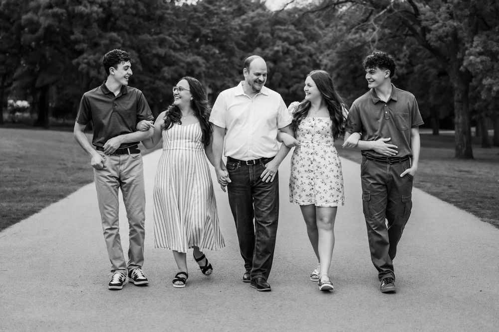 A group of five people walk together and laugh while strolling down a tree-lined path in black and white.