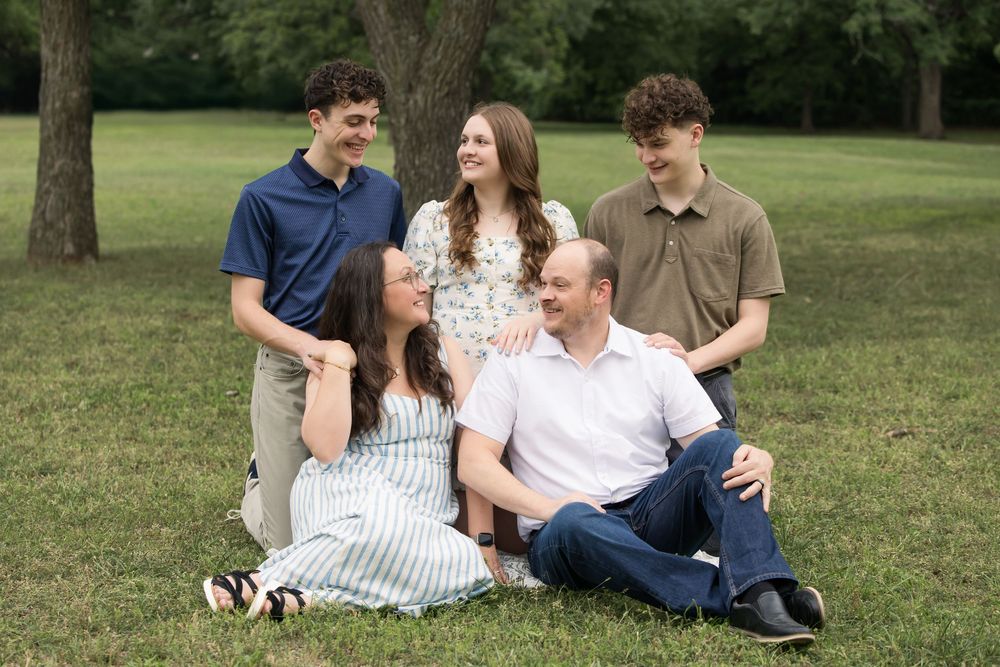A family group sits together on grass in a park setting during a casual outdoor photo session.