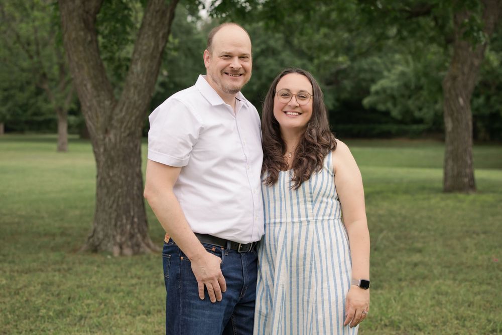 A couple stands close together outdoors on grass with trees in the background during a summer photo session.