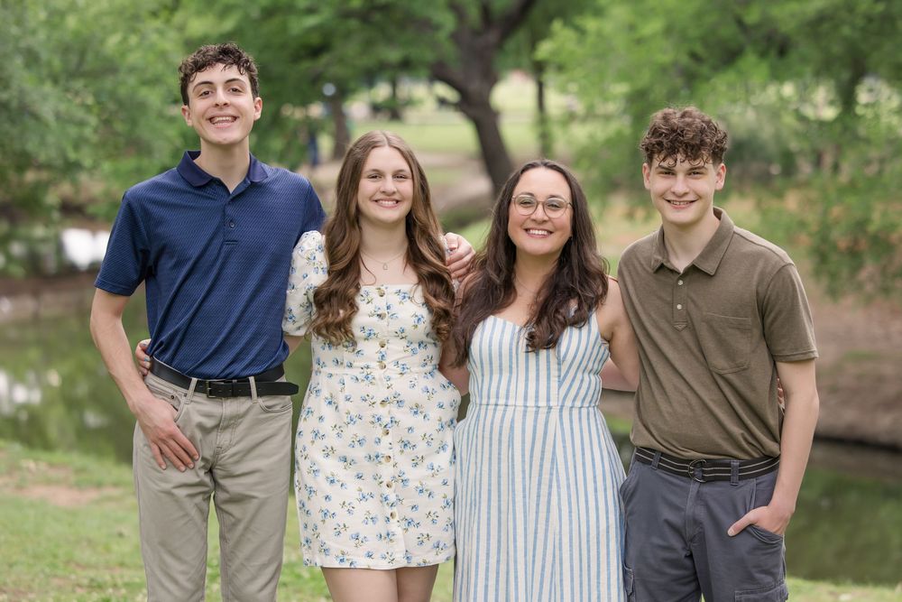 Four young people stand together outdoors in coordinated casual attire with a pond in the background.