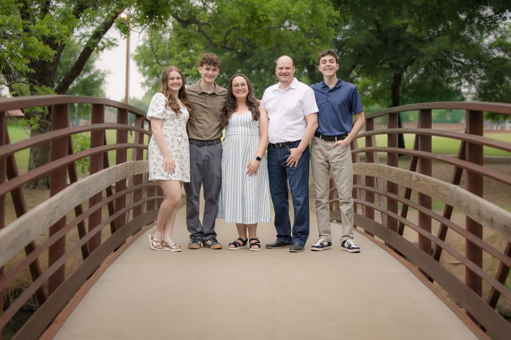 A group of five people stand together smiling on a wooden bridge surrounded by green trees.