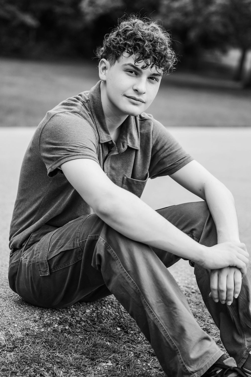 Black and white portrait of a young person with curly hair sitting outdoors in casual clothing.