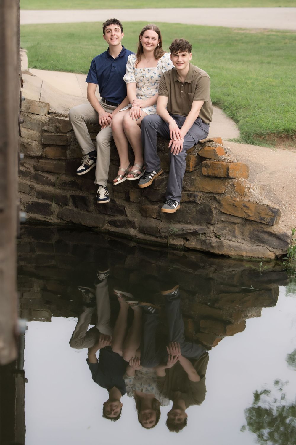Three people sit together on stone ledge above reflective water creating mirror-like image in park setting.