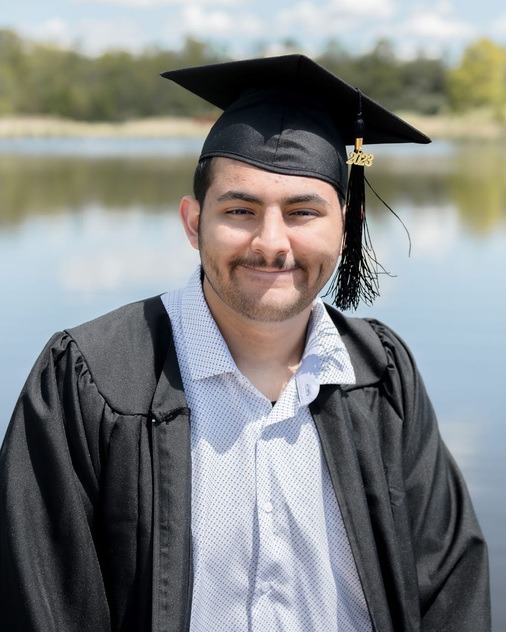 Graduate in cap and gown smiles for portrait with lake in background.