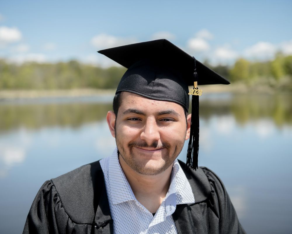 Graduate wearing traditional black cap and gown smiles in front of lake.
