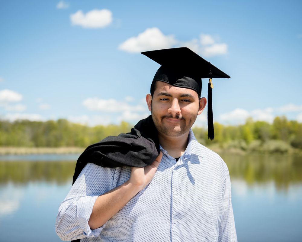 Graduate in cap and gown poses outdoors against blue sky and lake.
