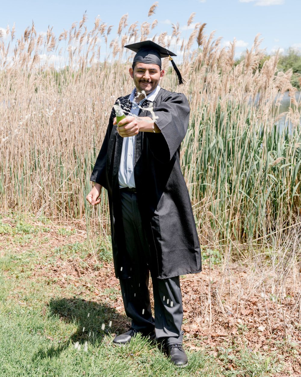 Graduate in full academic regalia stands among tall reeds holding diploma.