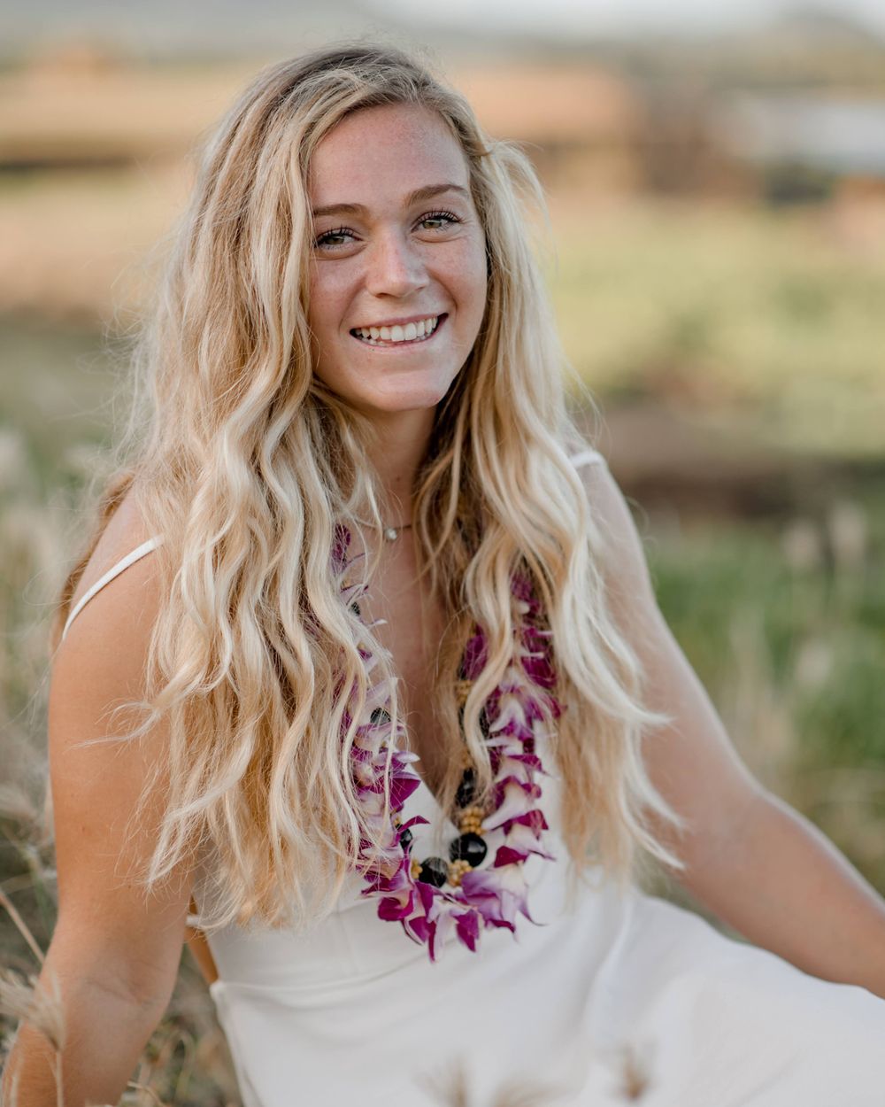 Woman in white dress with floral lei sits in outdoor field setting.
