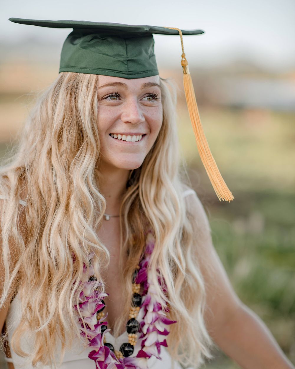 Graduate wearing green cap with gold tassel and purple flower lei photographed outdoors during sunset.