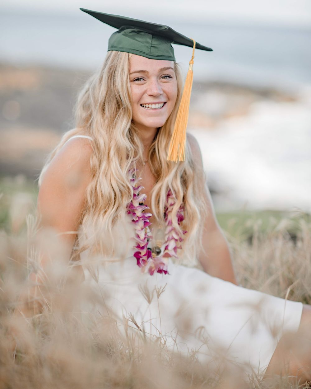 Graduation portrait with green mortarboard, gold tassel and purple lei against blurred natural background.