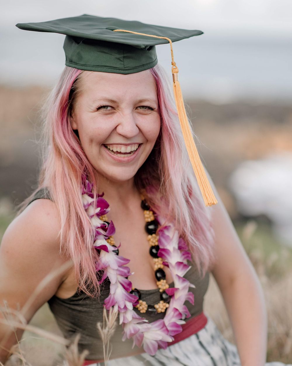 Graduate wearing green cap and floral lei with pink-tinted hair poses outdoors.