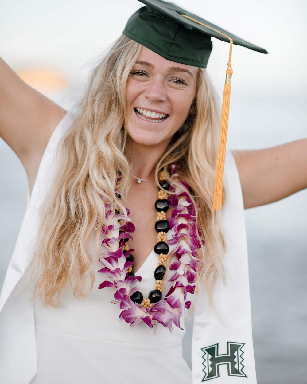Graduate in white dress with green cap and purple flower lei smiles with arms raised.