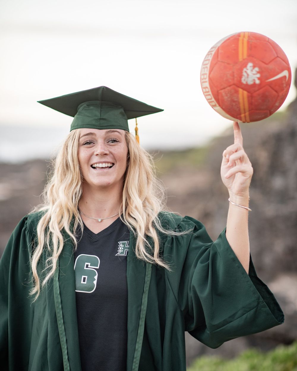 Graduate in green gown and cap spins basketball while celebrating outdoors.