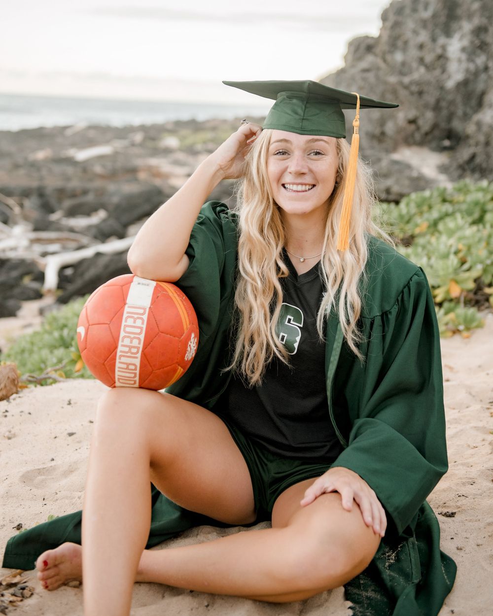 Graduate in green academic regalia poses with volleyball on coastal cliff.