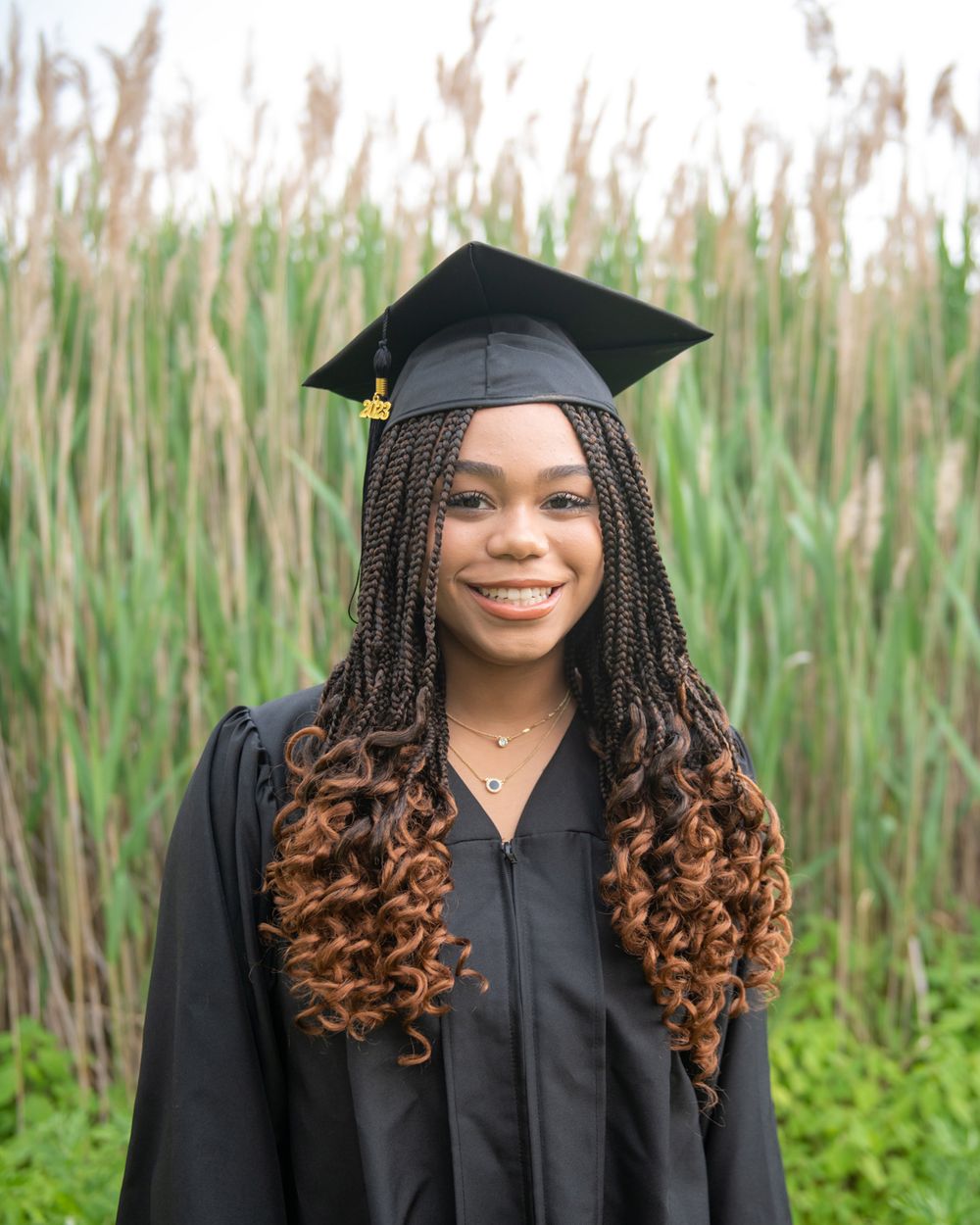 Graduate with braided hair and black cap and gown poses in front of reed grass.
