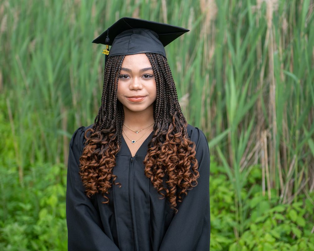 Graduate in black cap and gown stands in front of tall green reeds.