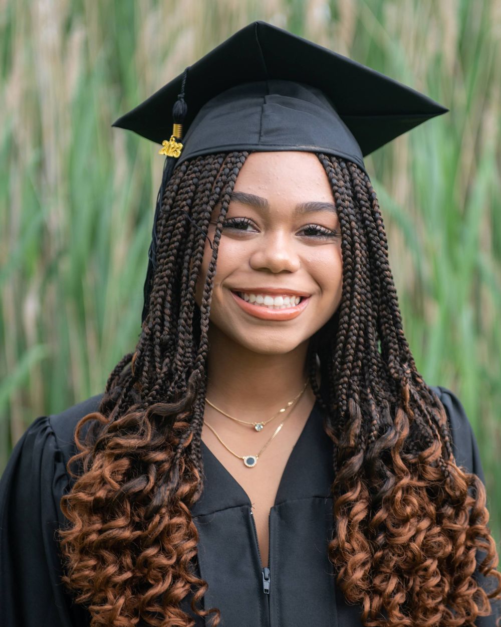 Graduate with long braided ombre hair poses in black cap and gown against natural background.