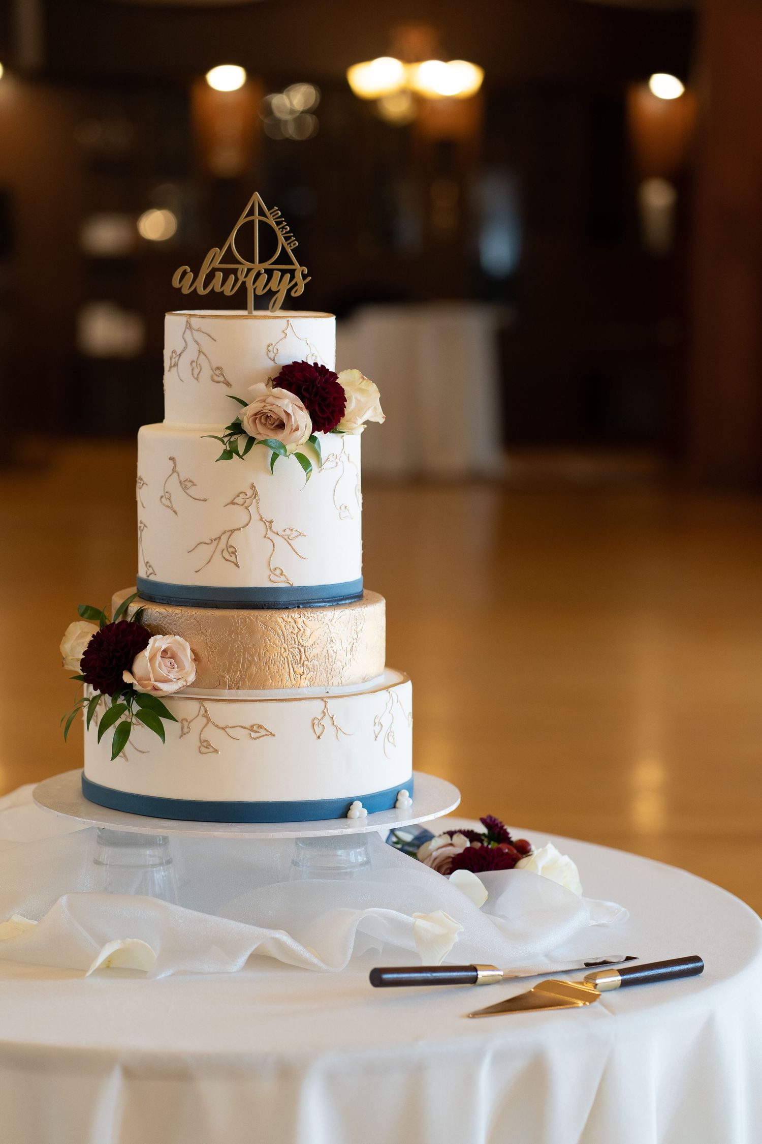 Elegant wedding cake with navy accents, gold geometric topper, and fresh roses on a white tablecloth.