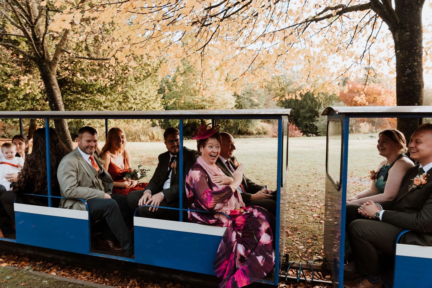 Wedding guests enjoy a ride on a blue and white outdoor train during a fall day with autumn leaves in the background.