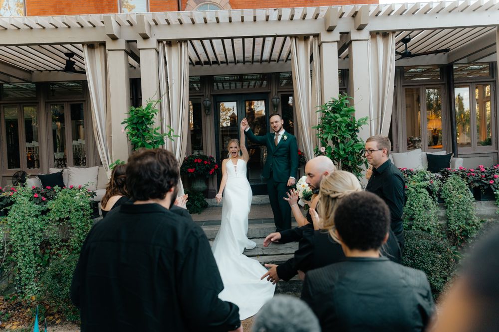 An outdoor wedding ceremony under a pergola surrounded by greenery and guests seated on wooden chairs.