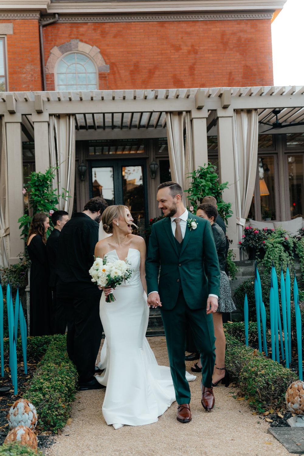 A couple walks together along a garden path outside a brick building with white columns during a wedding celebration.