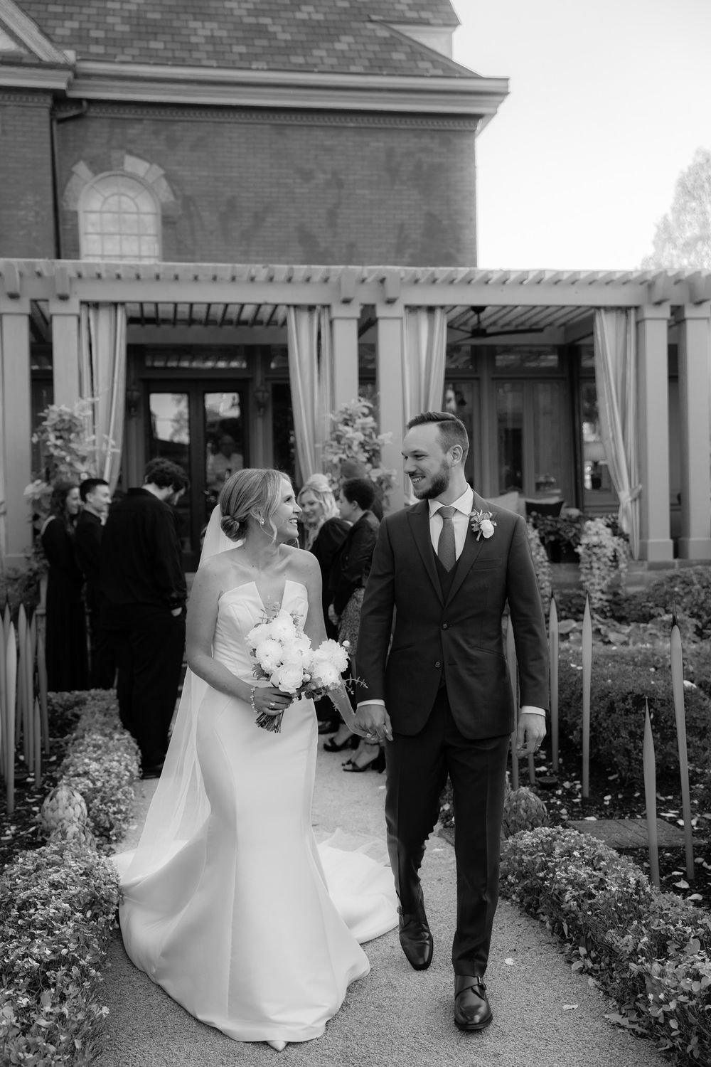 Newlyweds walk together along garden path outside historic venue after wedding ceremony in black and white photograph.
