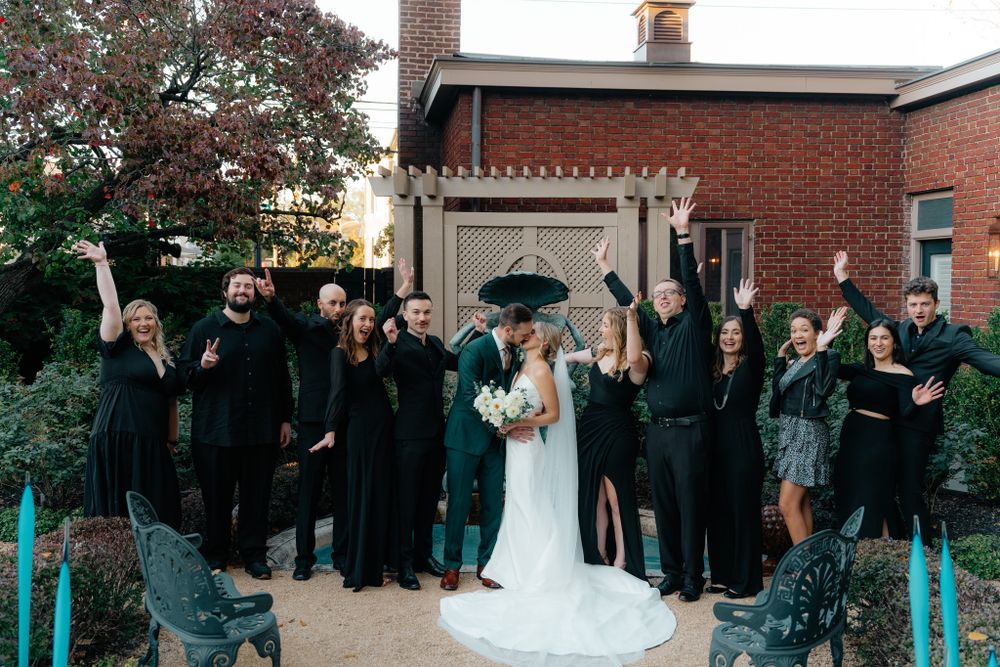 Wedding party dressed in black attire poses together with a couple under white pergola at brick venue.