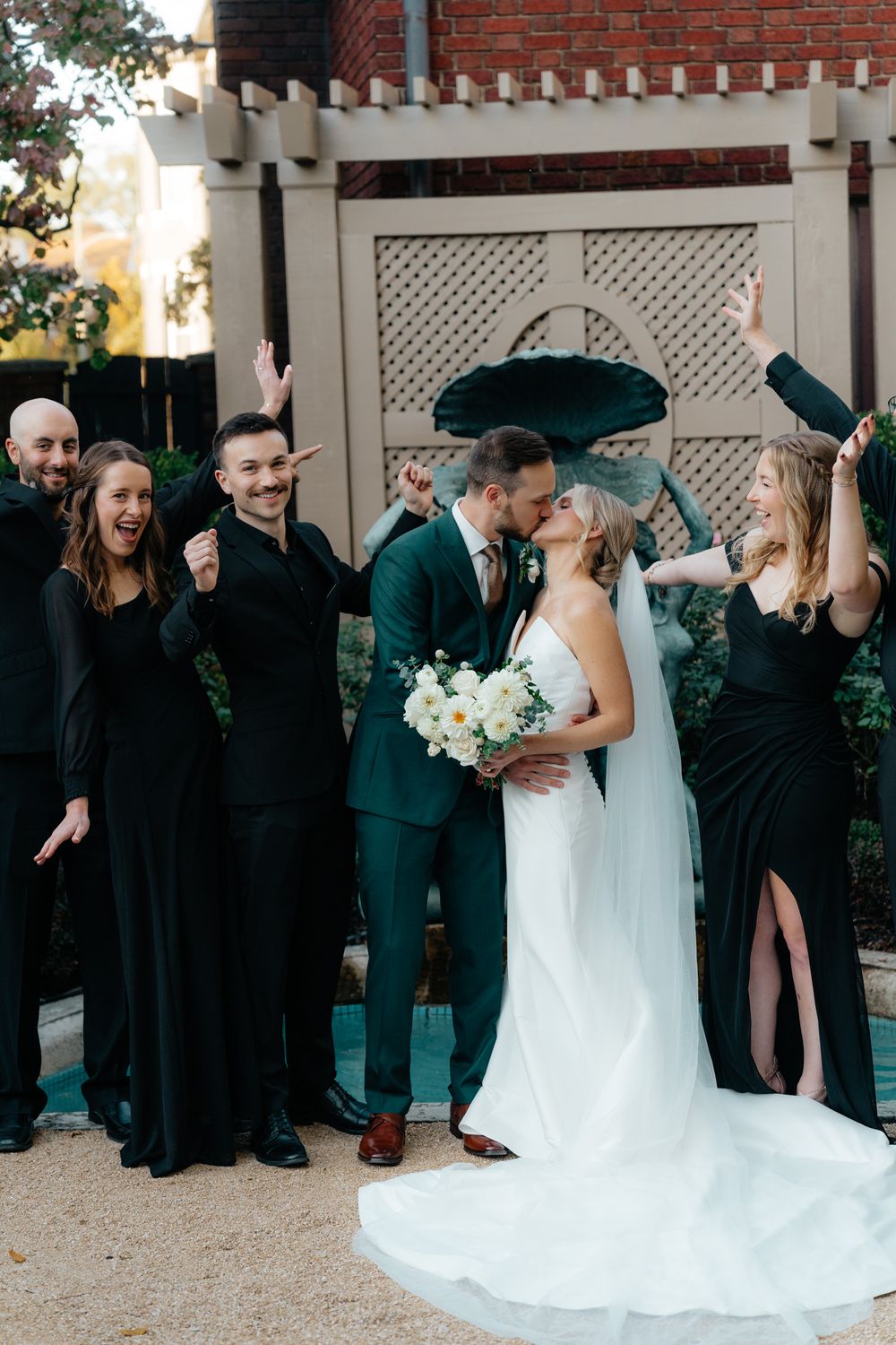 A joyful wedding party celebrates outside with the couple in emerald green and white attire with black bridesmaids dresses.