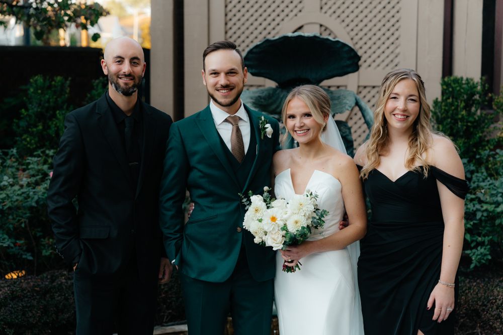 Wedding party stands outside venue with bride in white dress holding bouquet and groom in emerald green suit.