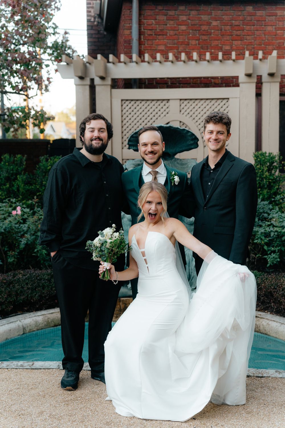 Wedding party poses together by fountain with brick building and pergola in background.