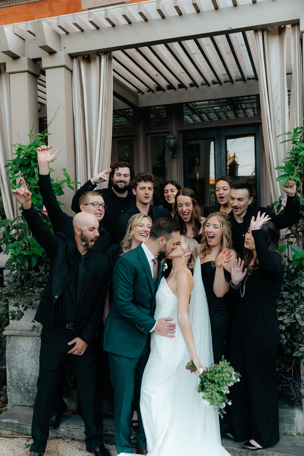 Wedding party celebrates with joyful expressions under a white pergola while dressed in black and teal formal attire.