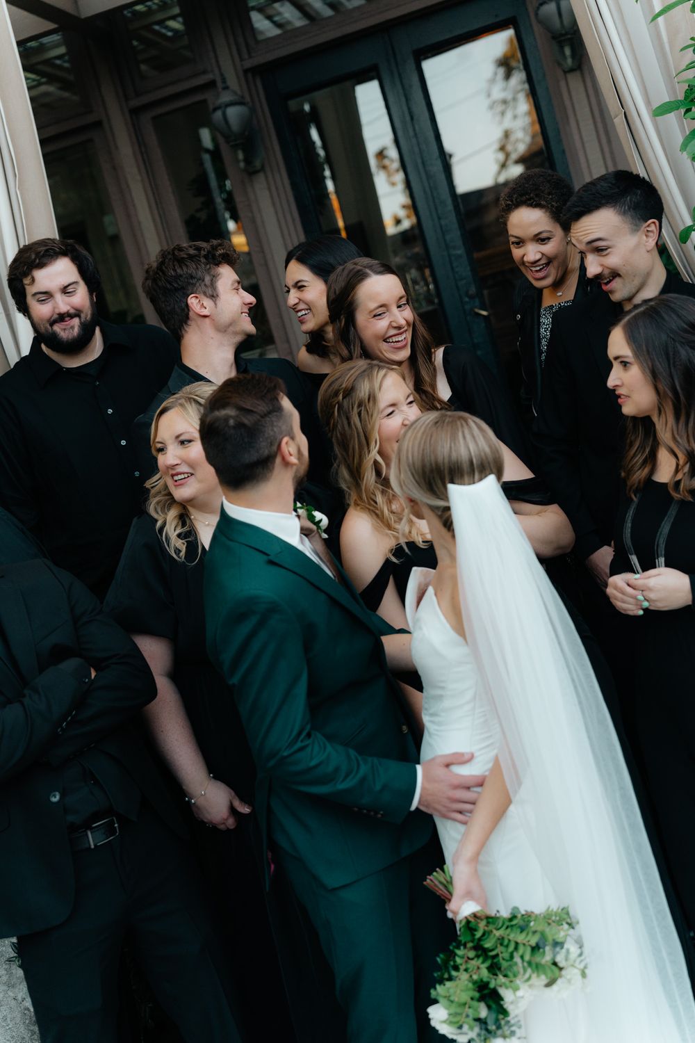 Wedding party shares a joyful moment together outdoors while bride in white gown holds green bouquet.