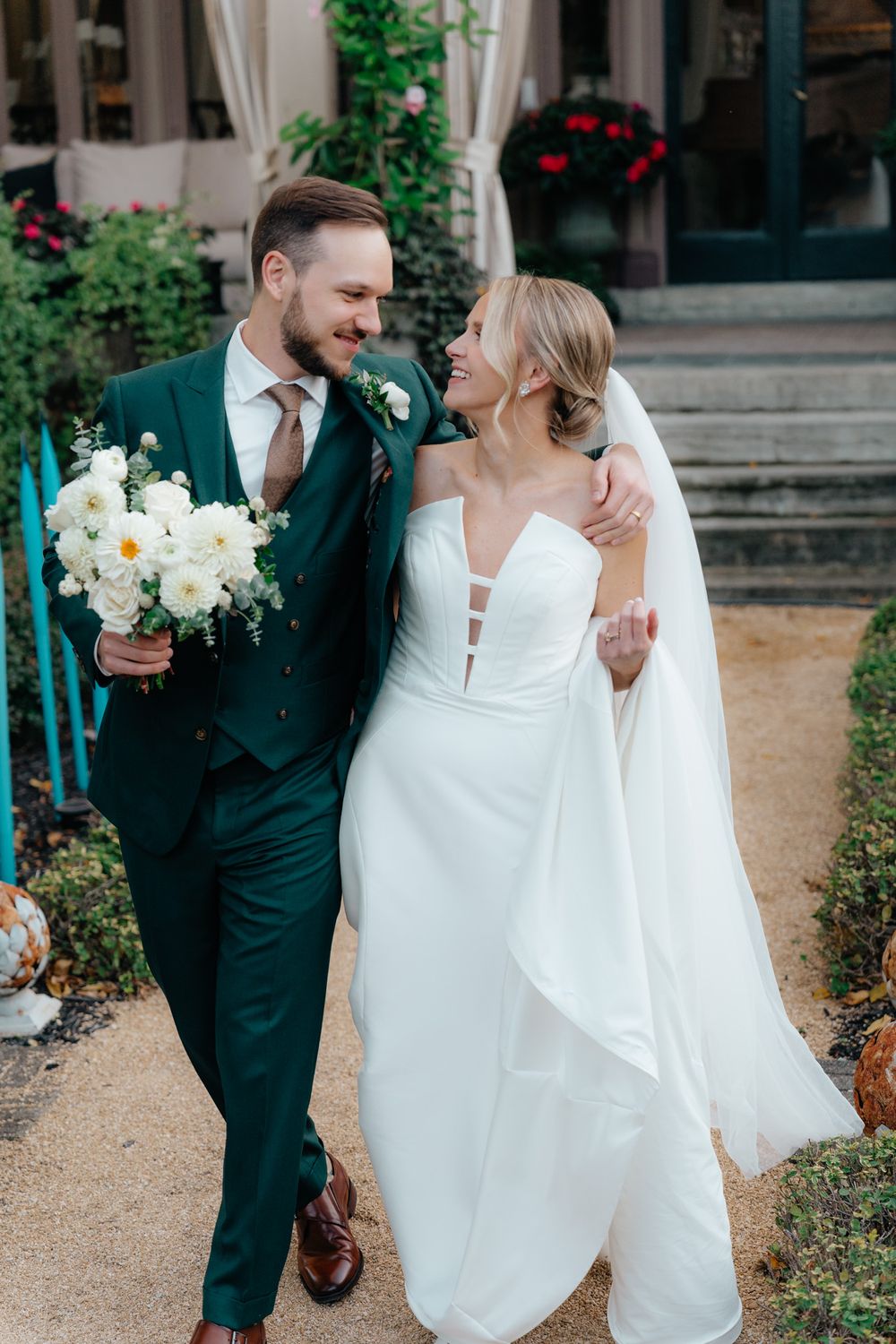 A couple in wedding attire walking together, with the bride in white and groom in emerald green suit holding a bouquet.
