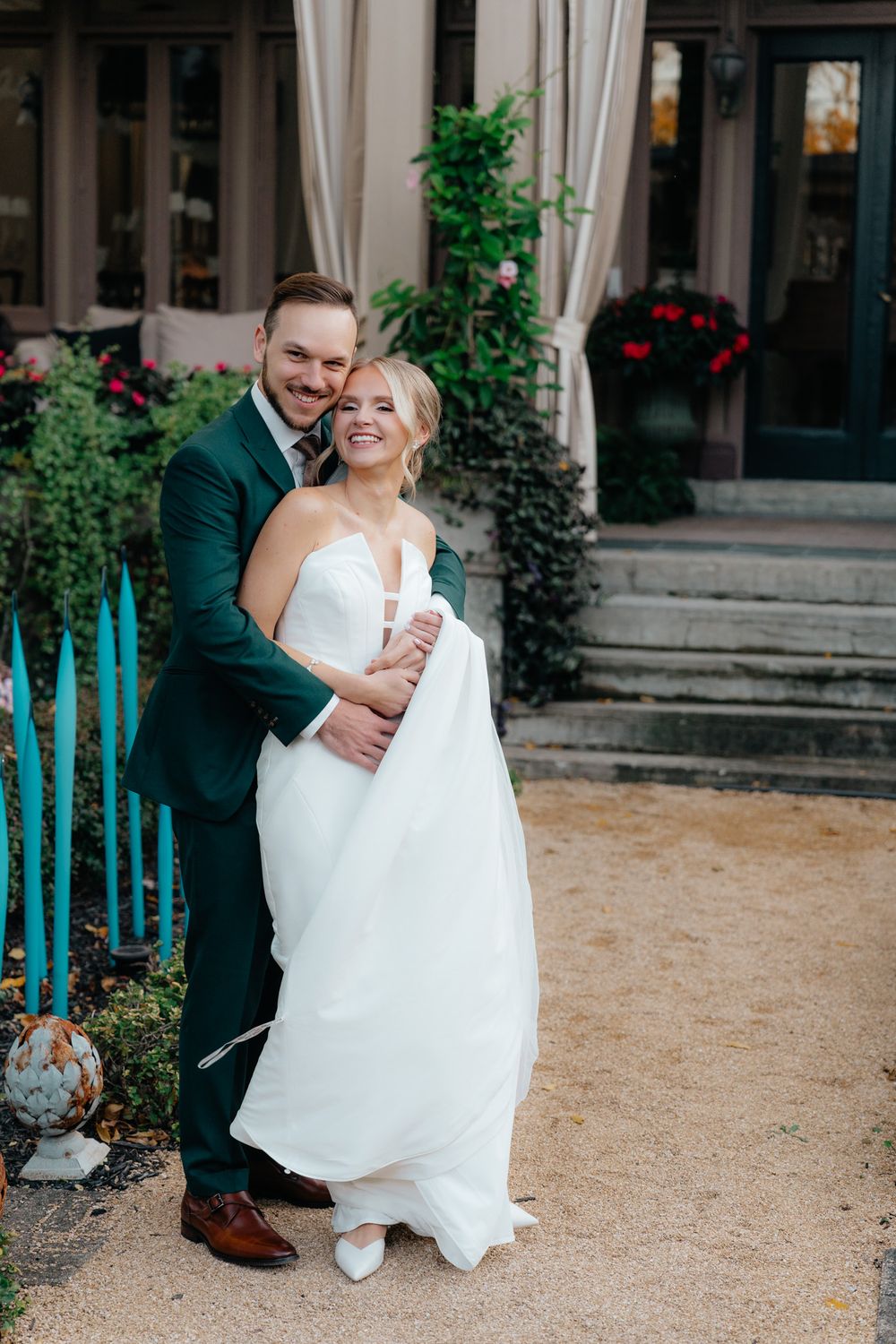 A bride in a white wedding dress and groom in dark green suit embrace on garden path with flowers and greenery around.