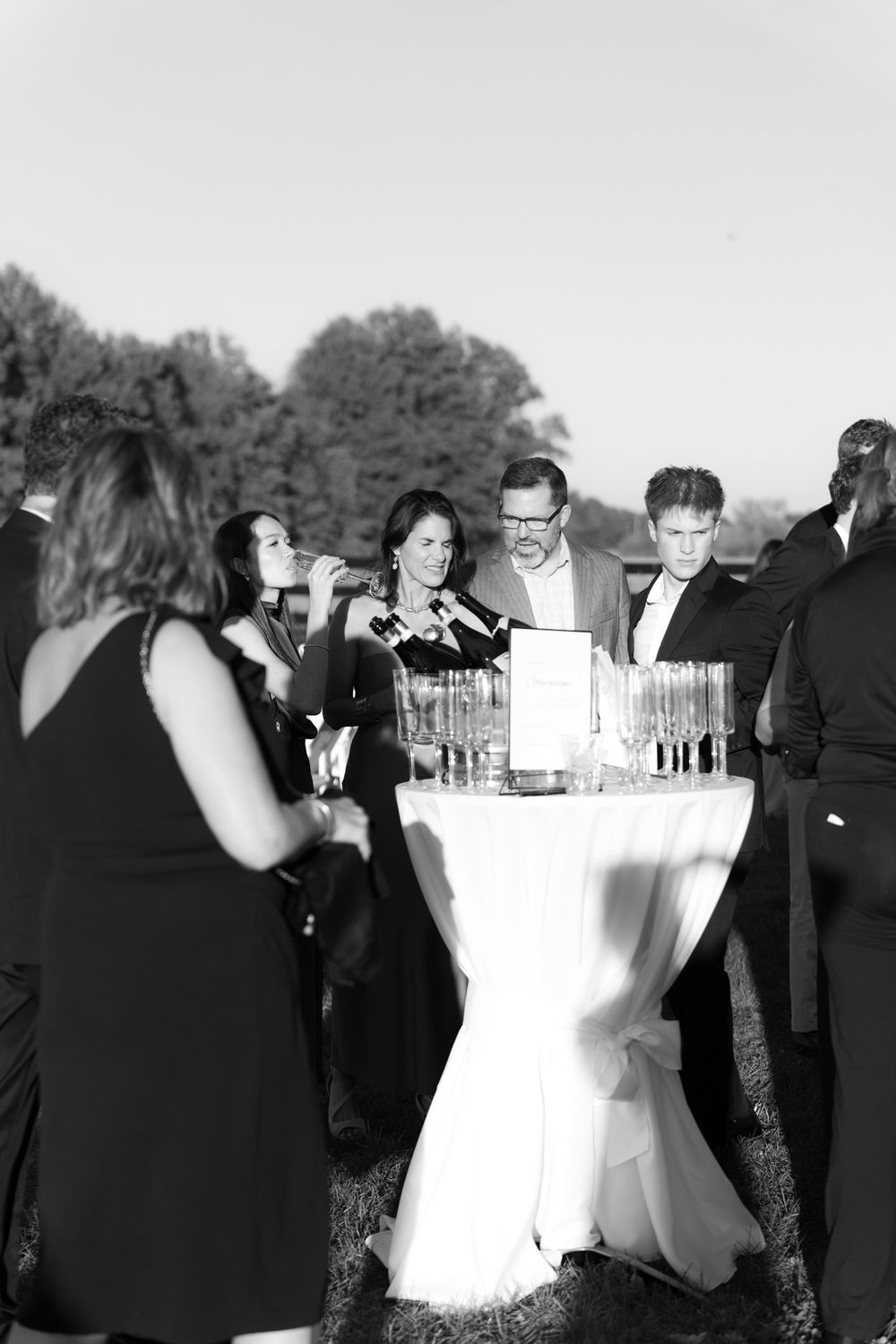 Wedding guests gather around a cocktail table with drinks at an elegant outdoor reception in black and white.