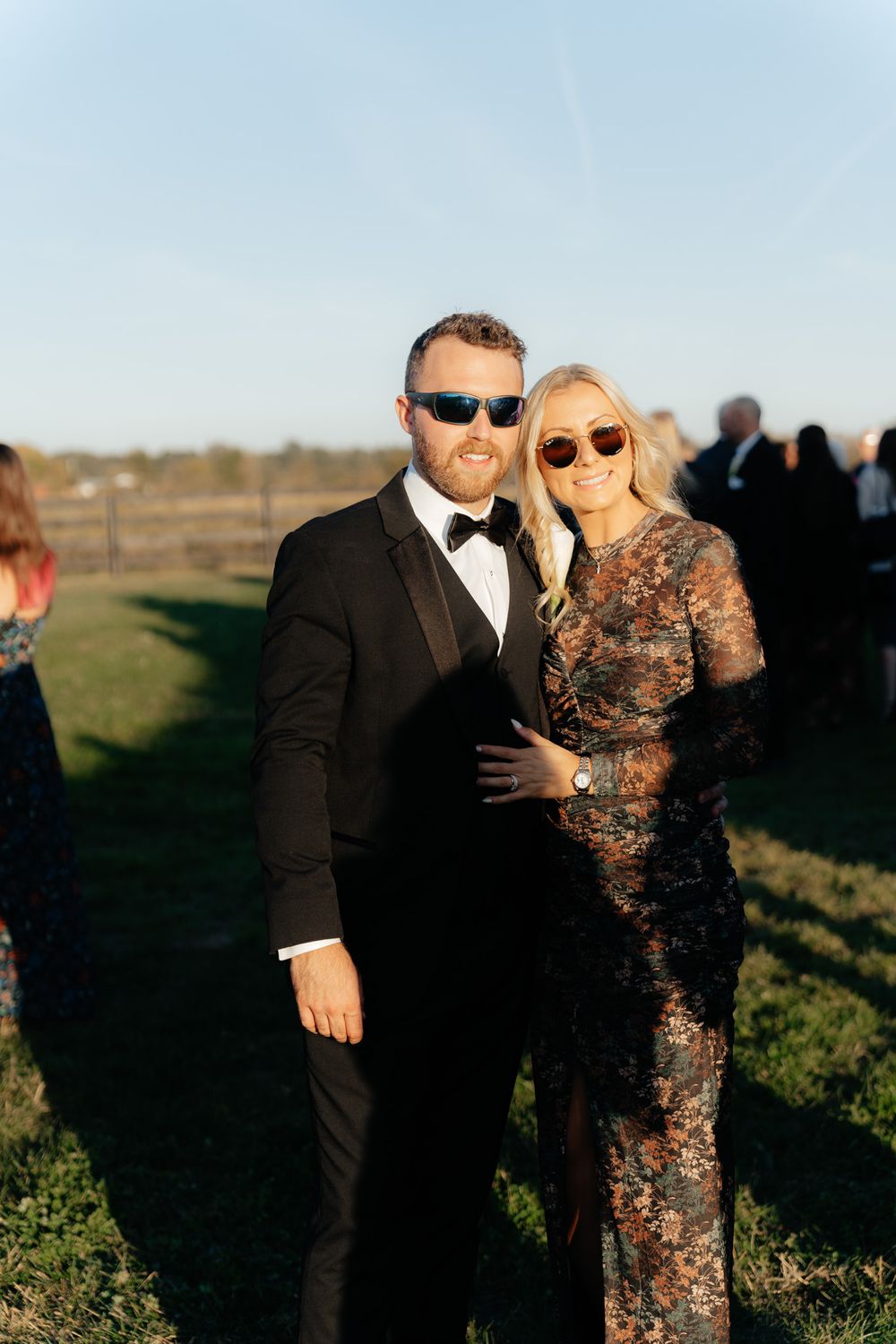 A couple in formal attire poses together at an outdoor event during golden hour.