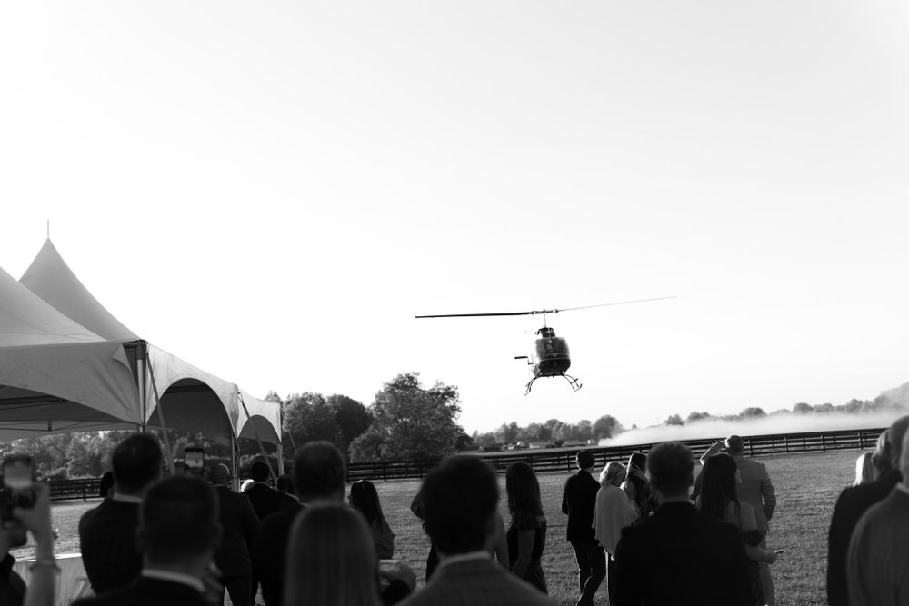 A silhouetted crowd watches a helicopter flying overhead at an outdoor event in black and white photography.
