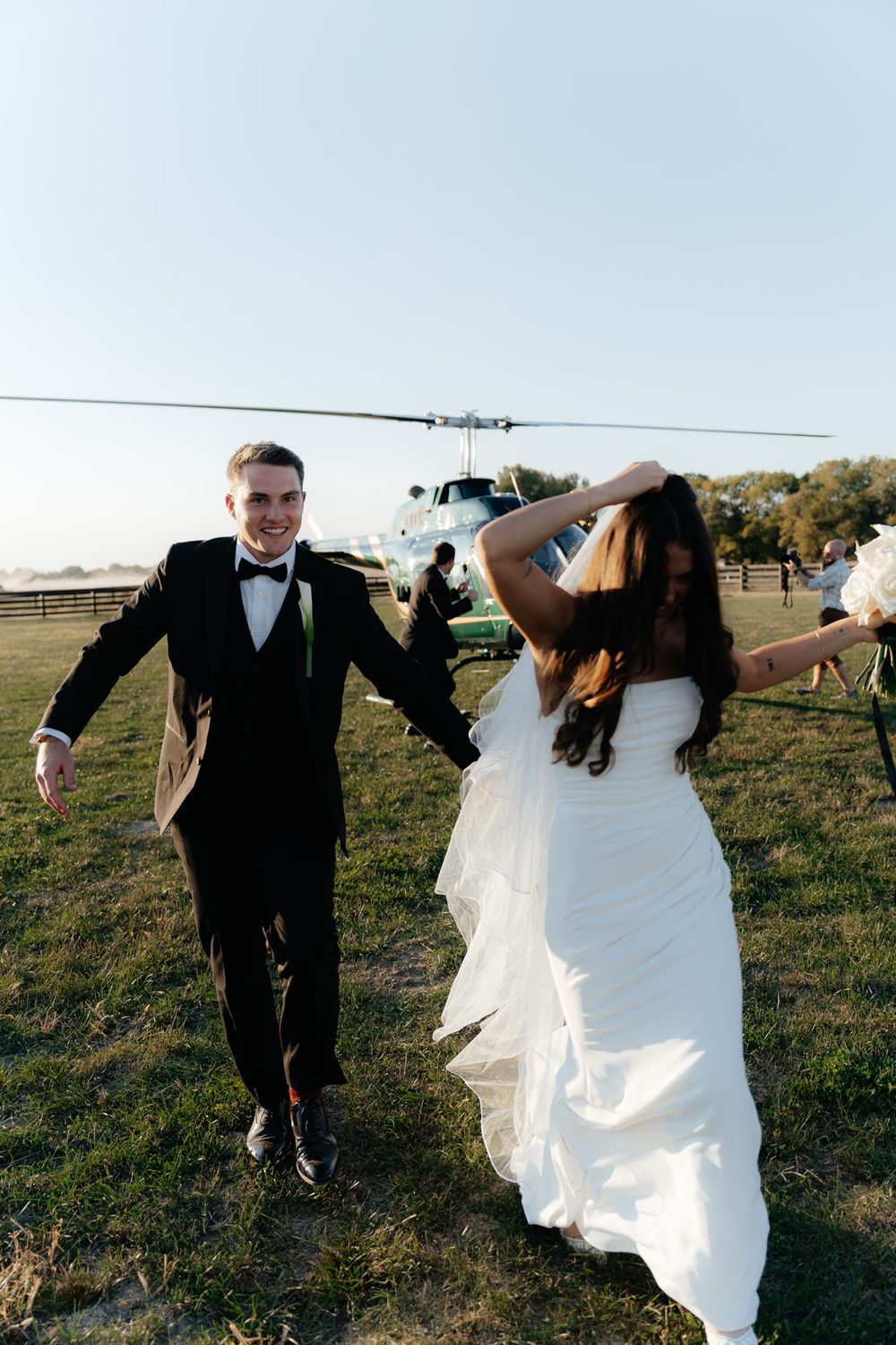 Newlyweds running together on grass with helicopter in background.