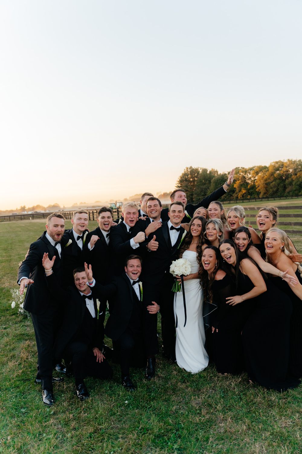 Wedding party in formal black attire poses together in a field at sunset.