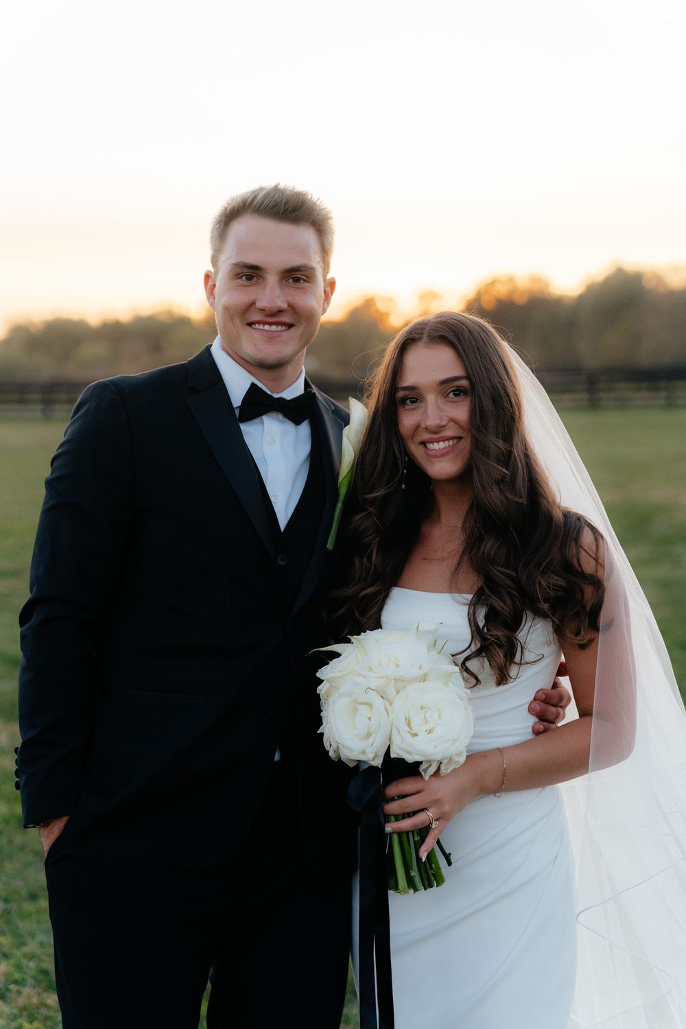 A couple in formal wedding attire poses outdoors at sunset with natural scenery in the background.