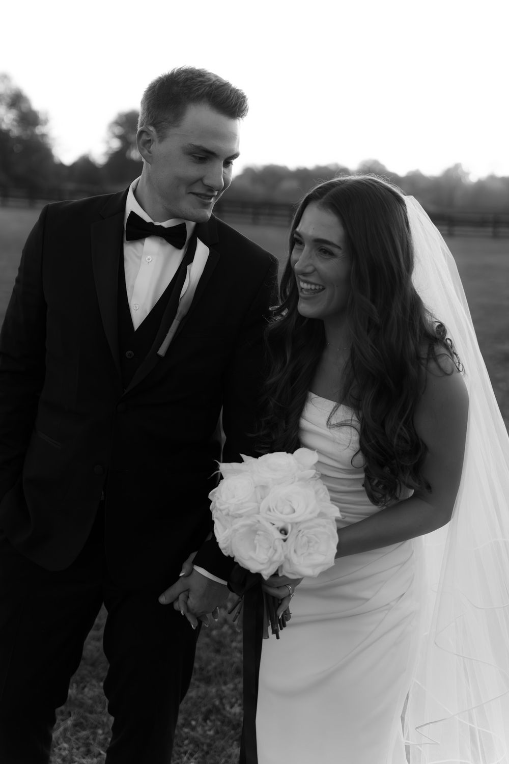 A black and white portrait of newlyweds sharing a joyful moment outdoors with white roses.